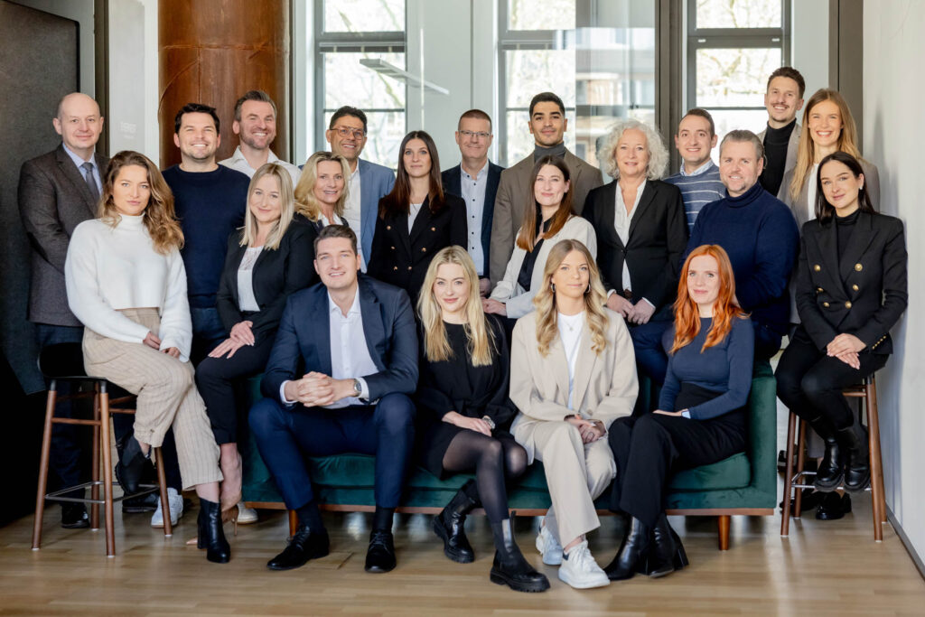 Teamfoto von 20 Leuten. Einige stehen, andere sitzen. Sie sind Business-casual gekleidet und lächeln.