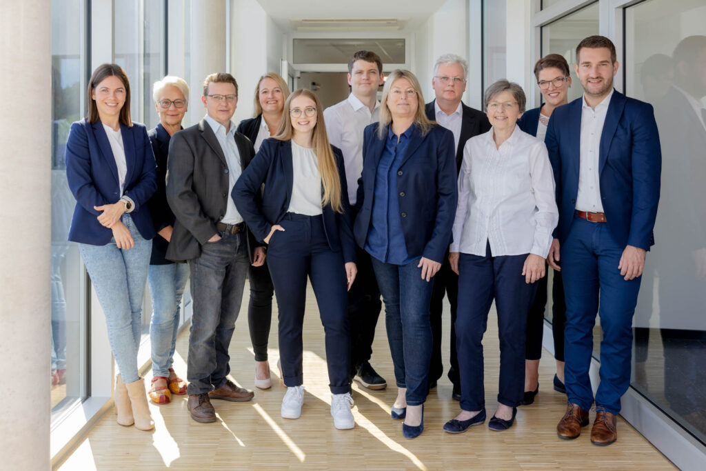Gruppenfoto von etwa 10 Personen in Business-Casual-Kleidung. Sie stehen in zwei Reihen und lächeln in die Kamera, vor einem neutralen Hintergrund. Das Bild vermittelt Professionalität und Teamgeist.