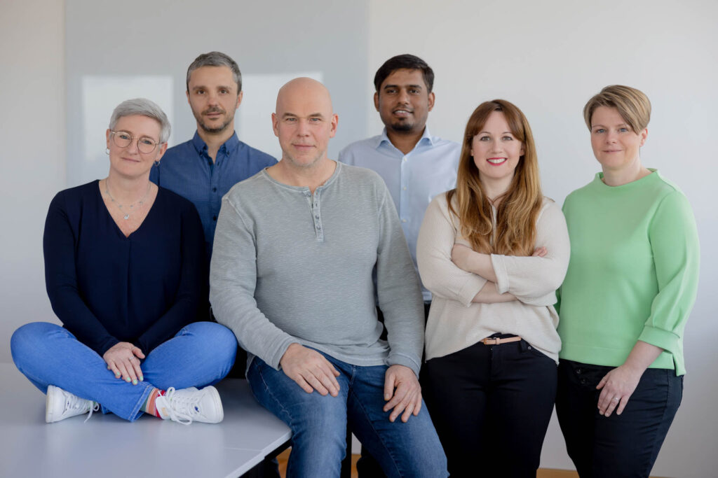Gruppenfoto von einem Team. Eine Frau sitzt im Schneidersitz auf einer Kommode. Alle lächeln.