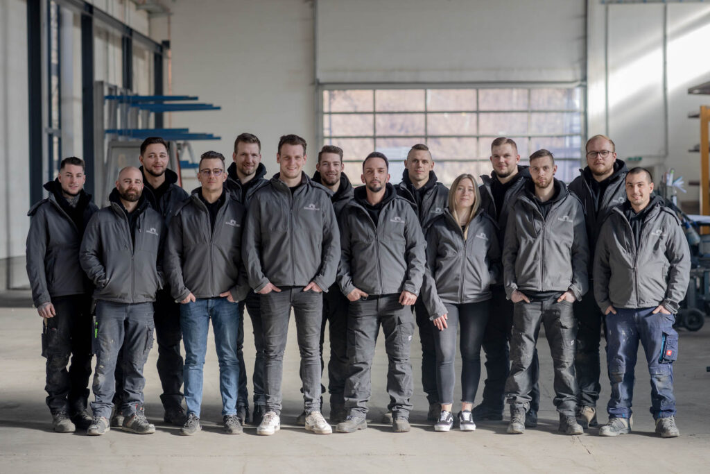 Teamfoto von Personen in Arbeitskleidung. Sie haben sie Hände in den Taschen. Sie lächeln und wirken sympathisch.