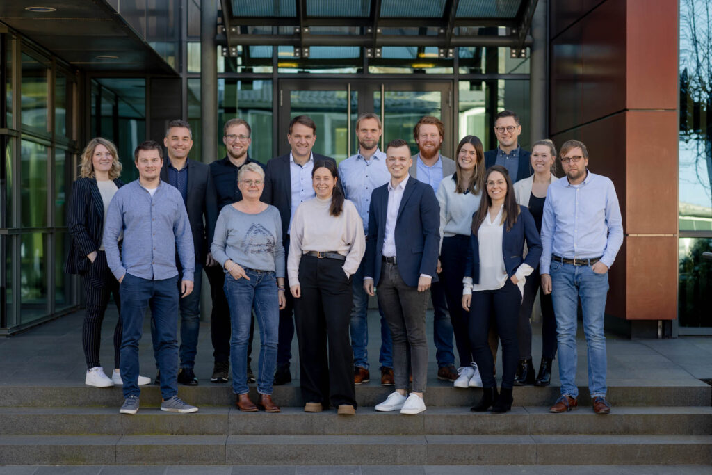 Teamfoto mit circa 20 Personen. Sie stehen in zwei Reihen vor einem Gebäude und lächeln.