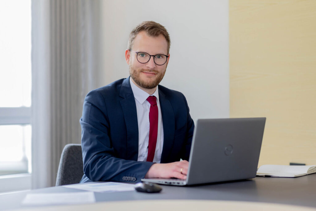 Vermögensberater bei der Volksbank. Er trägt einen Anzug, eine rote Krawatte und hat einen Laptop vor sich.