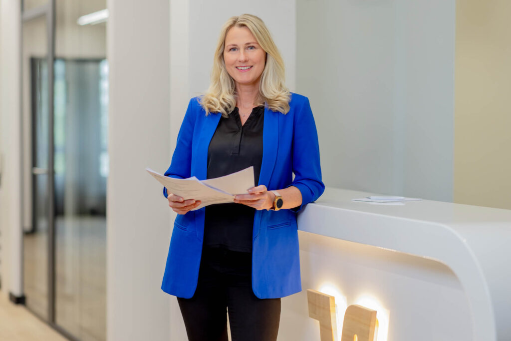 Vermögensberaterin bei der Volksbank. Sie trägt einen blauen Blazer und hält Unterlagen in der Hand. Sie lacht.