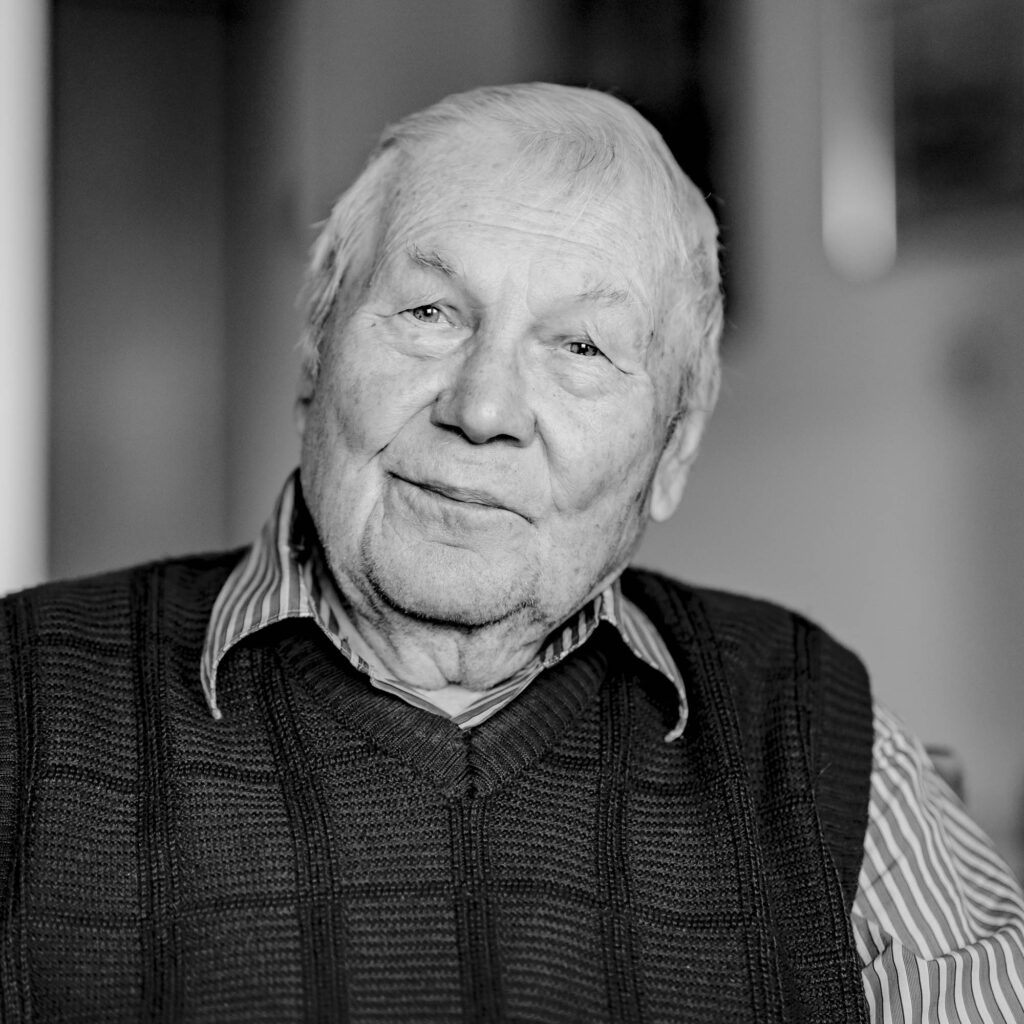 Schwarz-weiß Portrait eines älteren Mannes mit Hemd um Pullover. Er lächelt.
