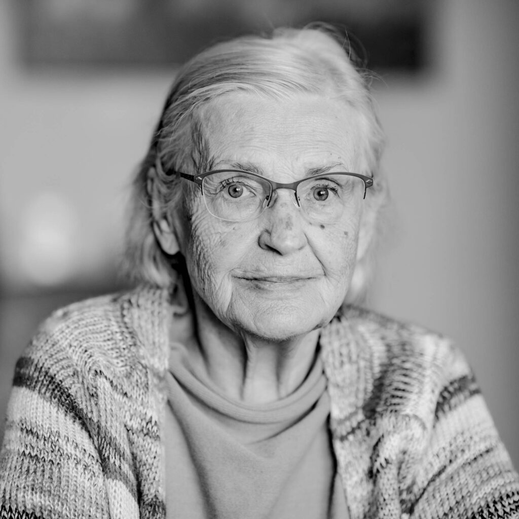 Schwarz-weiß Portrait einer älteren Frau mit schulterlangen Haaren und Brille. Sie lächelt.