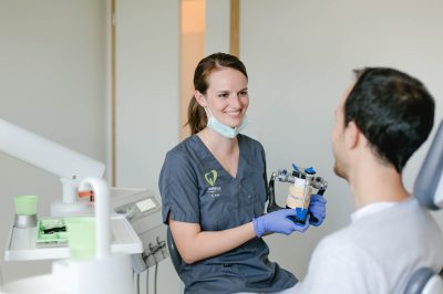 Eine Zahnärztin hält ein Modell in den Handy. Vor ihr sitzt ein Patient.